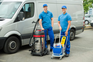 Two,Happy,Male,Cleaners,Standing,With,Cleaning,Equipments,In,Front